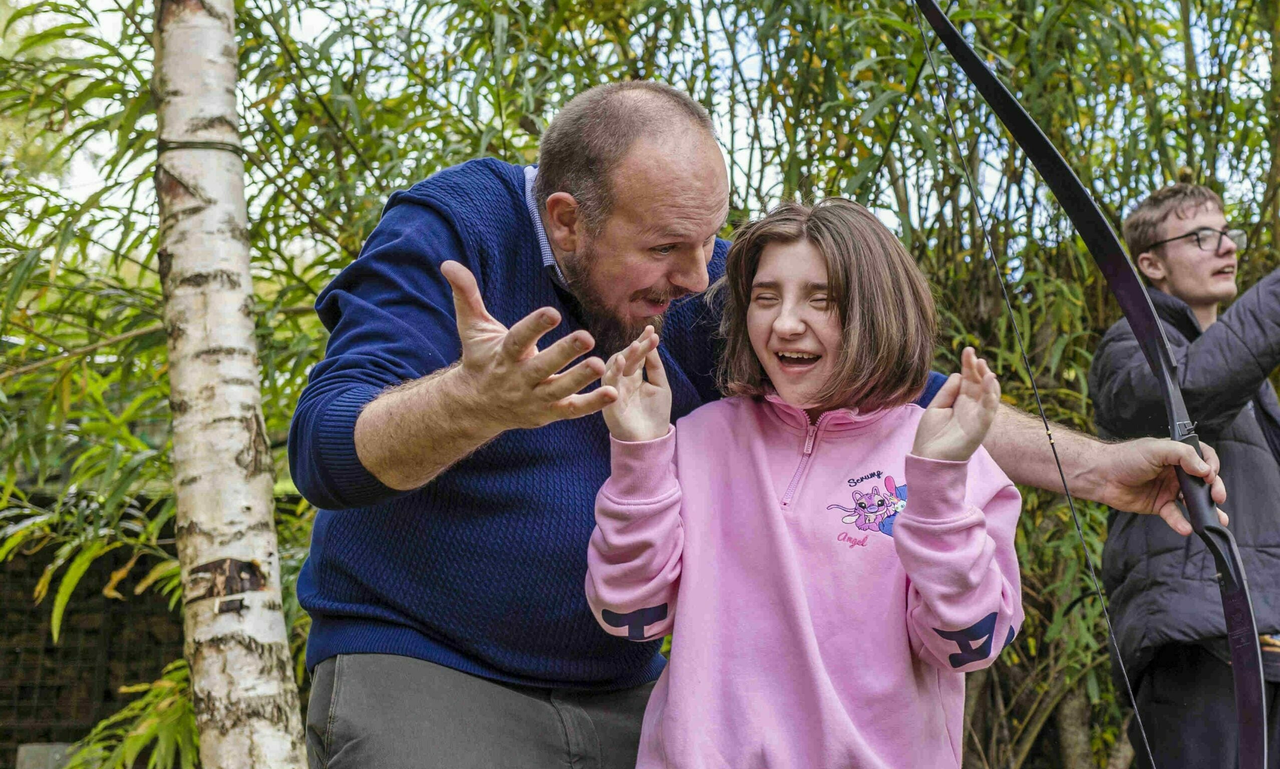 A man and a young girl are engaged in an archery lesson outdoors. The man, wearing a blue sweater, is demonstrating how to hold a bow while the girl, in a pink hoodie, looks excited and slightly surprised. In the background, another person is preparing to shoot an arrow. Lush greenery surrounds the scene, creating a natural setting.