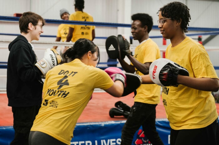 A group of young people in yellow t-shirts engaged in a boxing training session in a gym. One person is wearing pink boxing gloves and practicing punches with another, while two others are smiling and interacting nearby. The gym features a boxing ring and training equipment in the background.