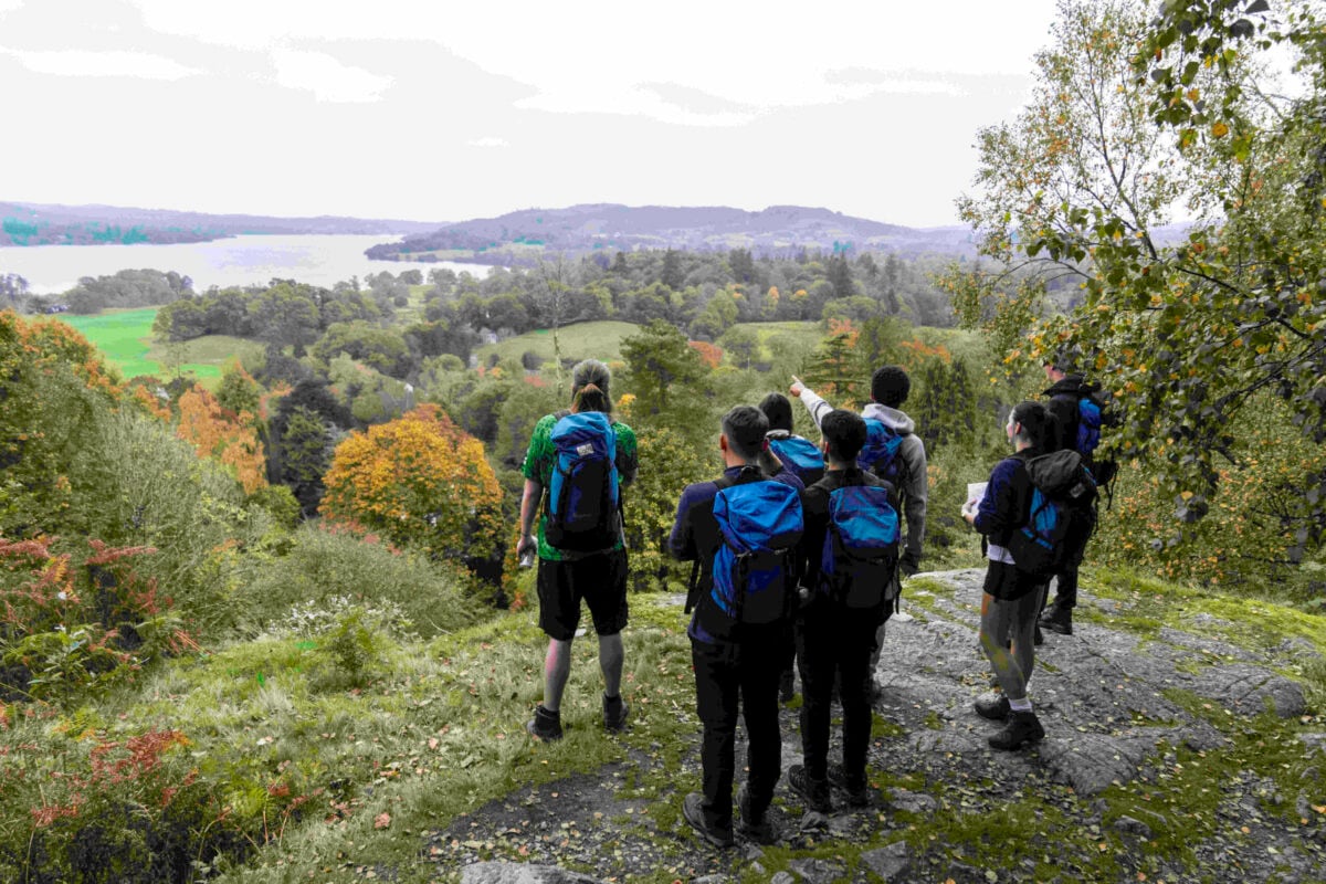 A group of six people, wearing backpacks, stands on a rocky outcrop overlooking a scenic landscape.