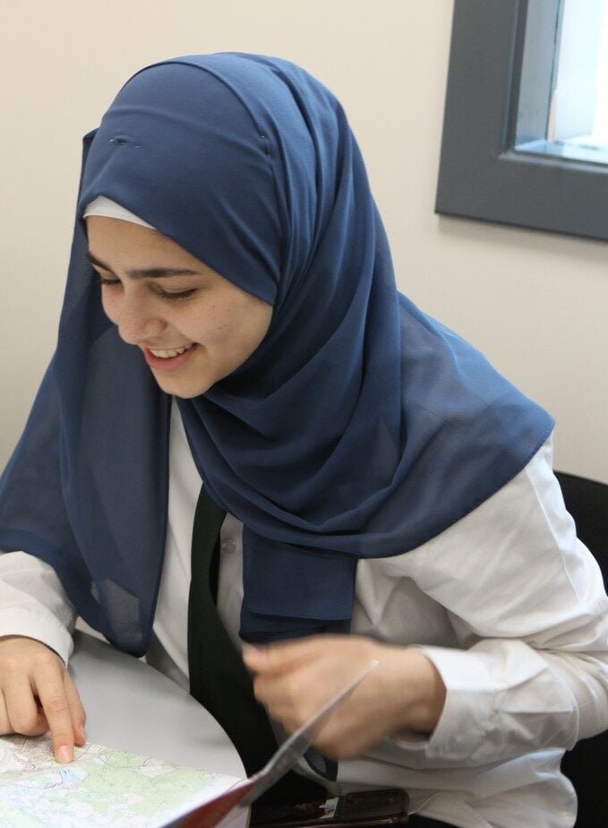 Person seated at a table, wearing a blue headscarf and white shirt, looking down at a printed map and holding a document.