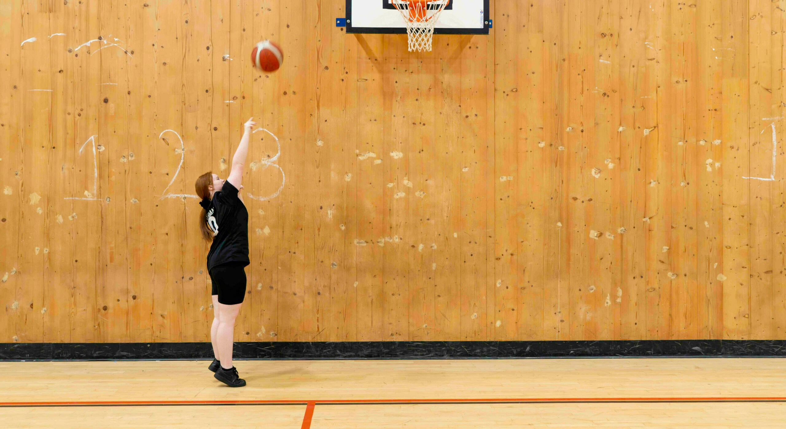 A young person standing under a basketball hoop after throwing a ball towards it.