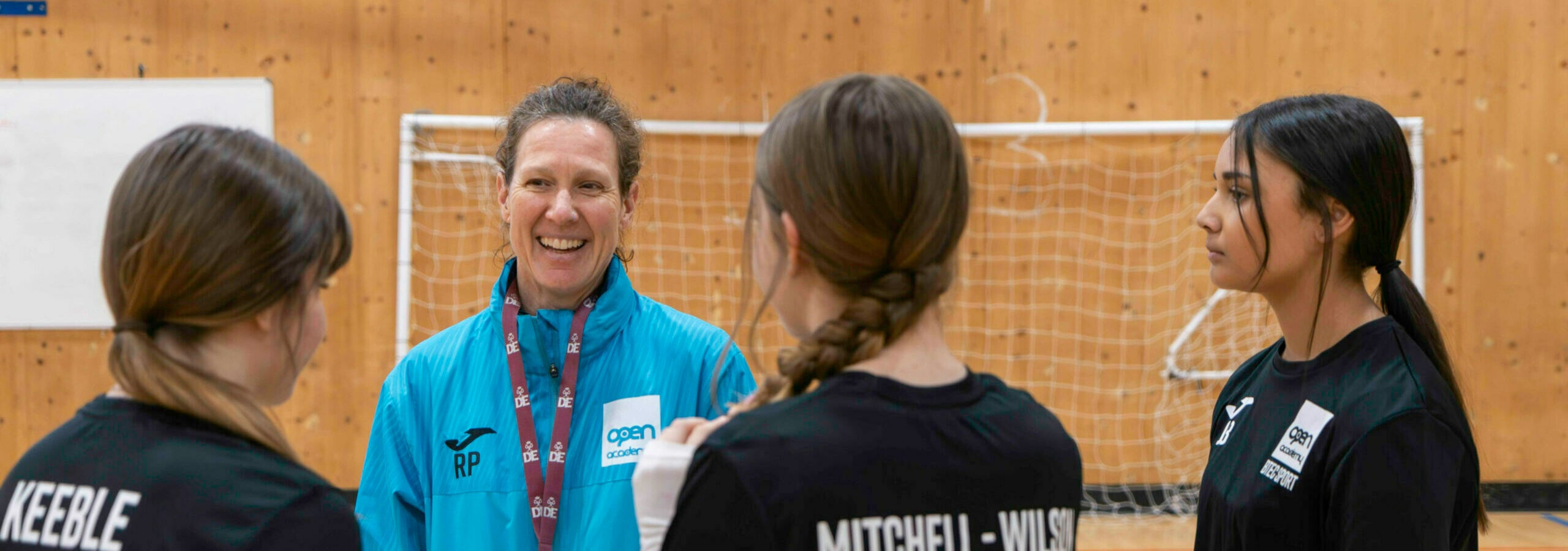 A P.E. teacher in a turquoise jacket talking to three pupils.