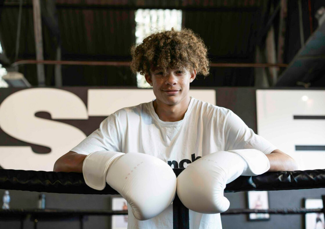 Harvey has curly brown hair, is wearing a white t-shirt and boxing gloves and leans on the edge of a boxing ring.