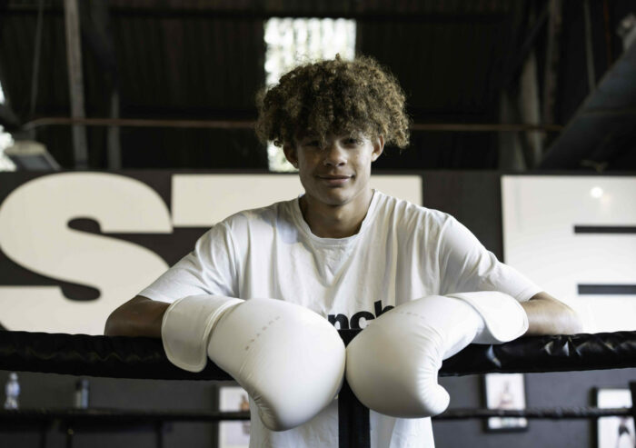Harvey has curly brown hair, is wearing a white t-shirt and boxing gloves and leans on the edge of a boxing ring.