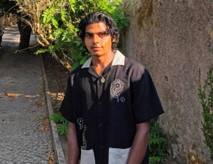 Ashwin stands on a sunlit outdoor path beside a stone wall and leafy plants. The person wears a black short-sleeved button-up shirt with a light-coloured collar, large light pockets, and white line illustrations on the front, along with a necklace. The path is paved with small stones and lined with trees and greenery.
