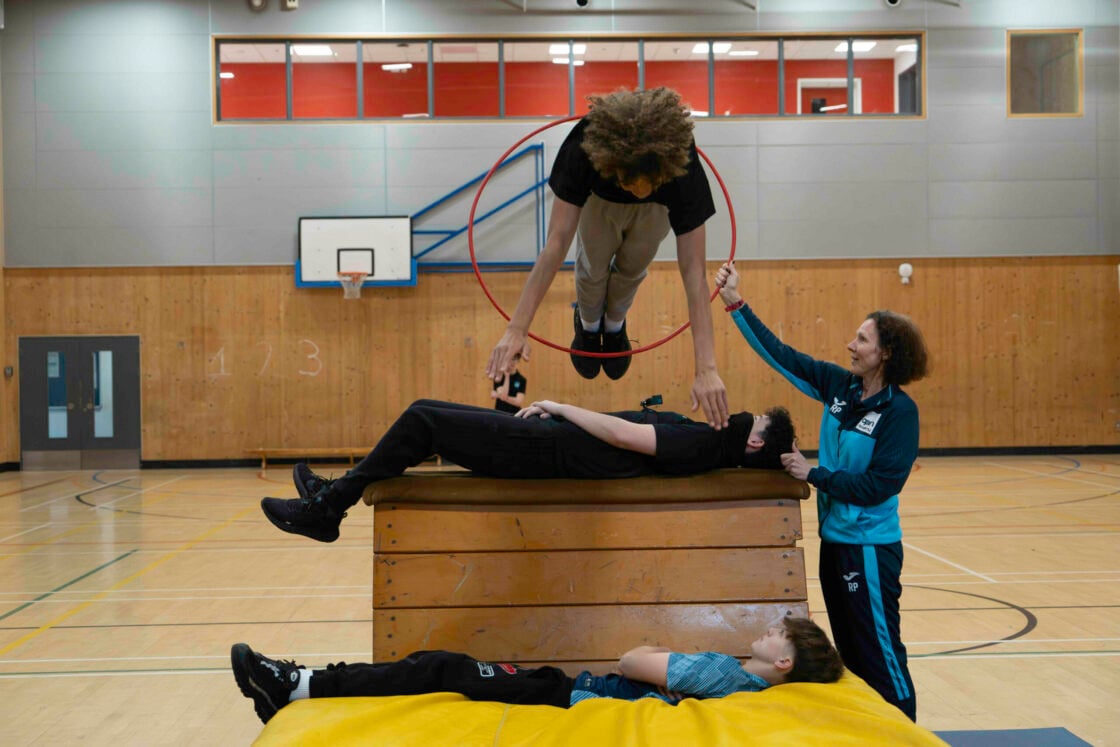 A young person jumps head-first through a hoop held by a teacher in a gym hall to practice circus skills for their Skills section.