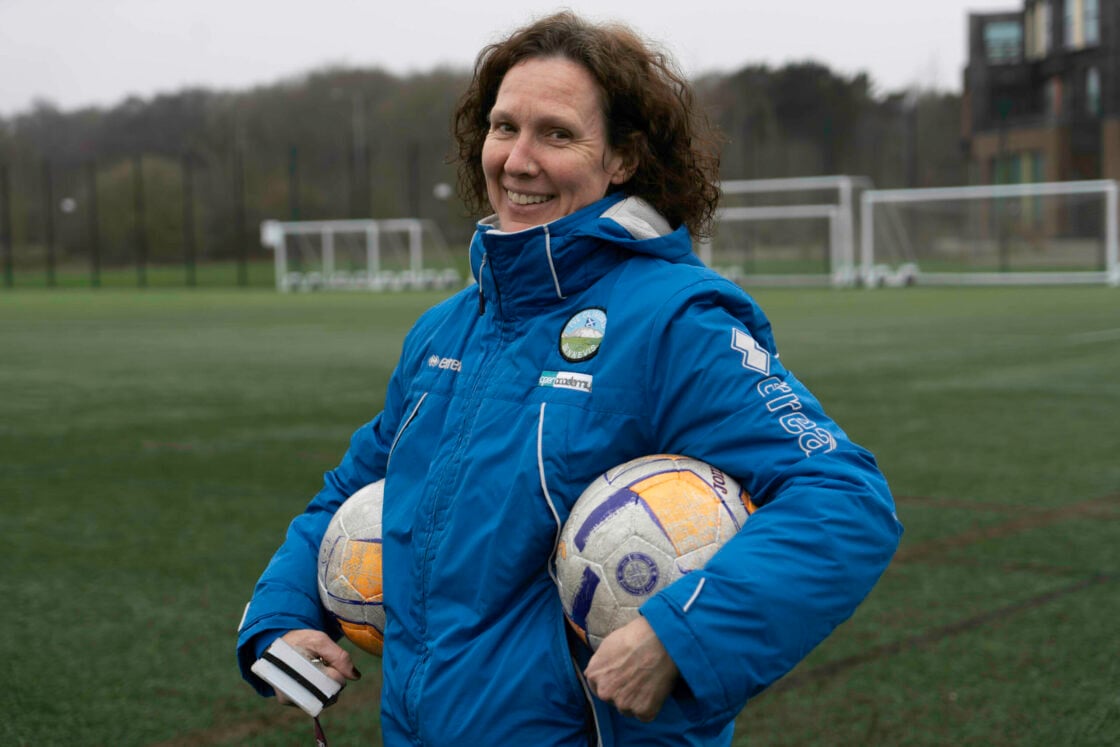 D of E Leader Ruth smiling on a football field wearing a blue coat and holding a football under each arm.