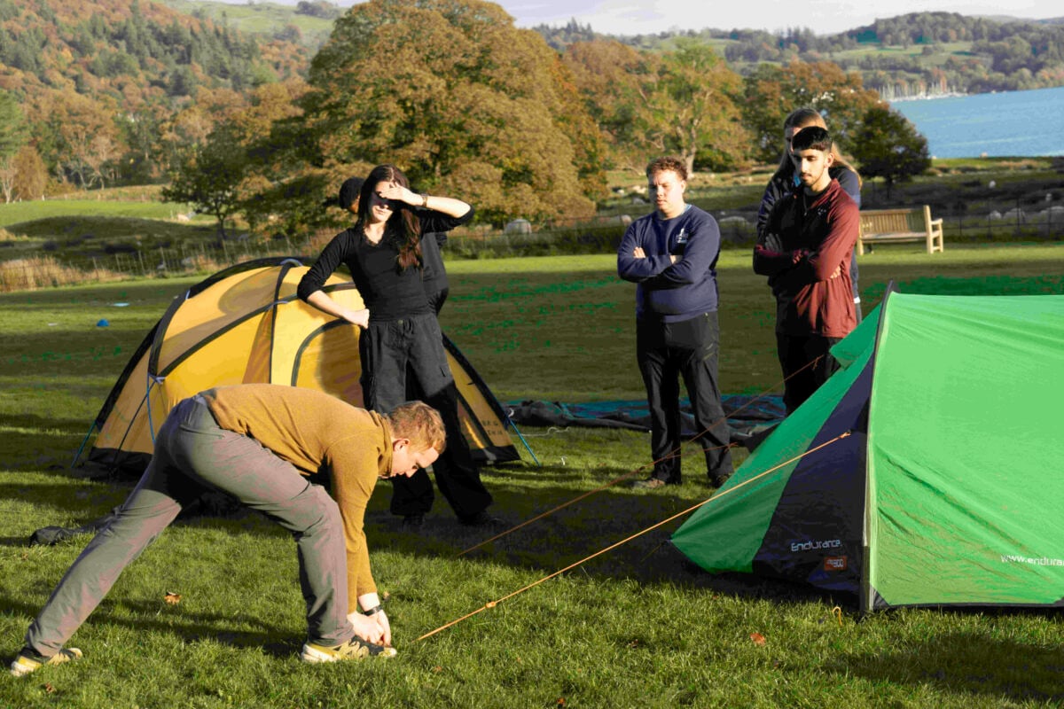 A D of E Leader shows three participants how to pitch a tent outside on a field.