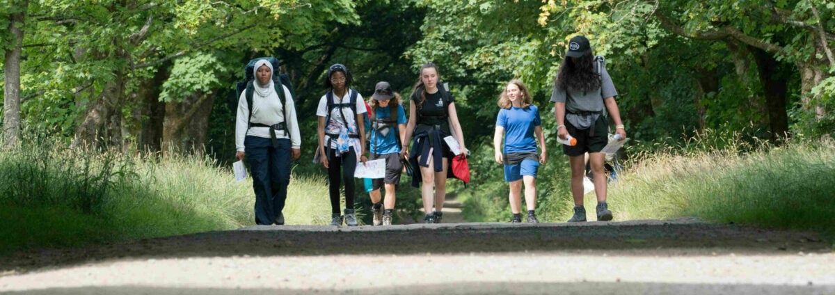 Six young people walking down a road lined with trees carrying rucksacks and holding maps.