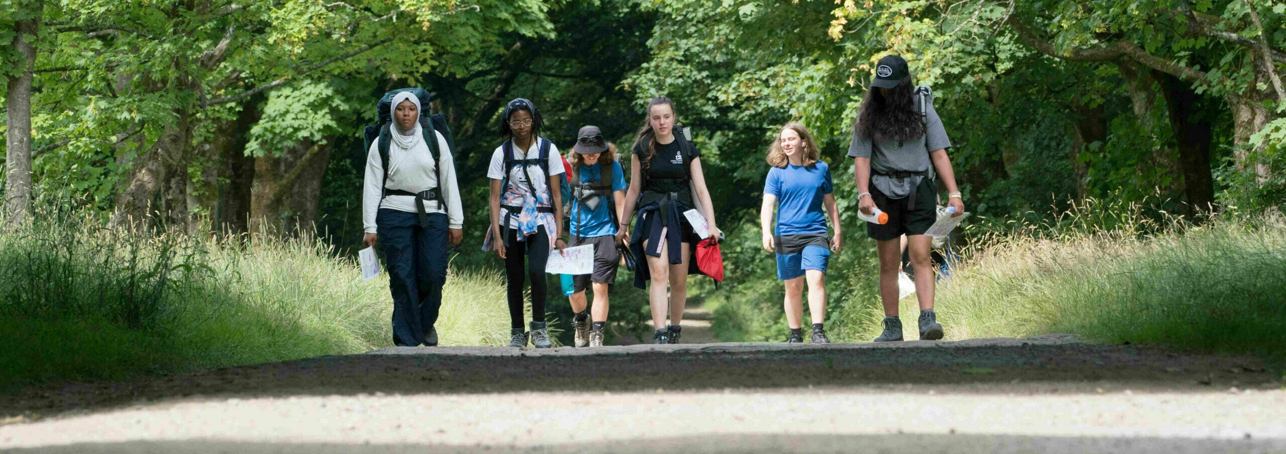 Six young people walking down a road lined with trees carrying rucksacks and holding maps.
