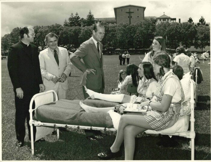 Black-and-white photograph of a group gathered on a grass field. In the centre, HRH The Duke of Edinburgh, Prince Philip, stands with one hand on their hip, speaking to several young people seated on and around a metal-framed bed placed outdoors. One young person lies on the bed with a leg in a plaster cast, while others sit nearby wearing striped uniforms. To the left, two adults in formal clothing, including one in clerical dress, stand observing. In the background, more people are scattered across the field, with a large building featuring a prominent cross and a row of trees behind it.