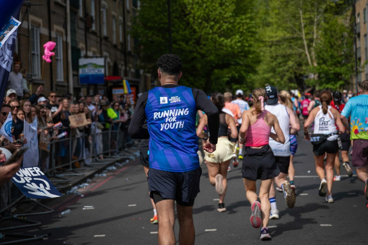 A group of runners move along a city street during a road race, viewed from behind. In the centre foreground, a runner wears a blue vest with white text reading “RUNNING FOR YOUTH” and a small “DofE Youth Without Limits” logo. Other runners in varied sportswear are ahead and to the sides. Crowds line the pavement behind barriers on the left, some holding signs and cheering. Trees and brick buildings frame the street in the background.