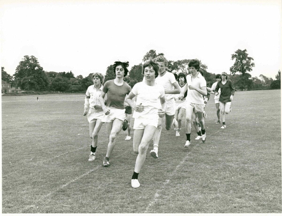 Black-and-white photograph of a group of young people running together across a wide, open grass field. The runners wear T-shirts, shorts and trainers, moving toward the camera in a loose cluster, with one runner slightly ahead in the centre. Trees and a few distant buildings line the background under an open sky, indicating an outdoor sports or cross-country setting.