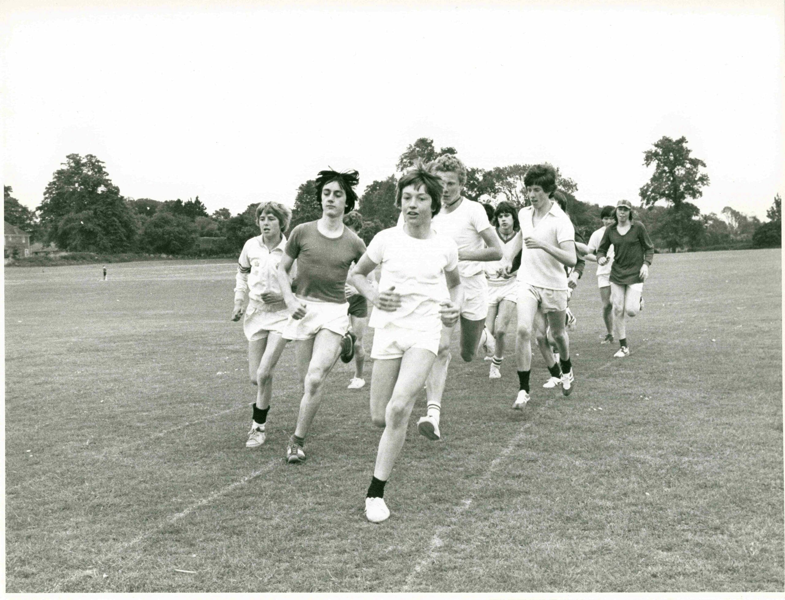 Black-and-white photograph of a group of young people running together across a wide, open grass field. The runners wear T-shirts, shorts and trainers, moving toward the camera in a loose cluster, with one runner slightly ahead in the centre. Trees and a few distant buildings line the background under an open sky, indicating an outdoor sports or cross-country setting.
