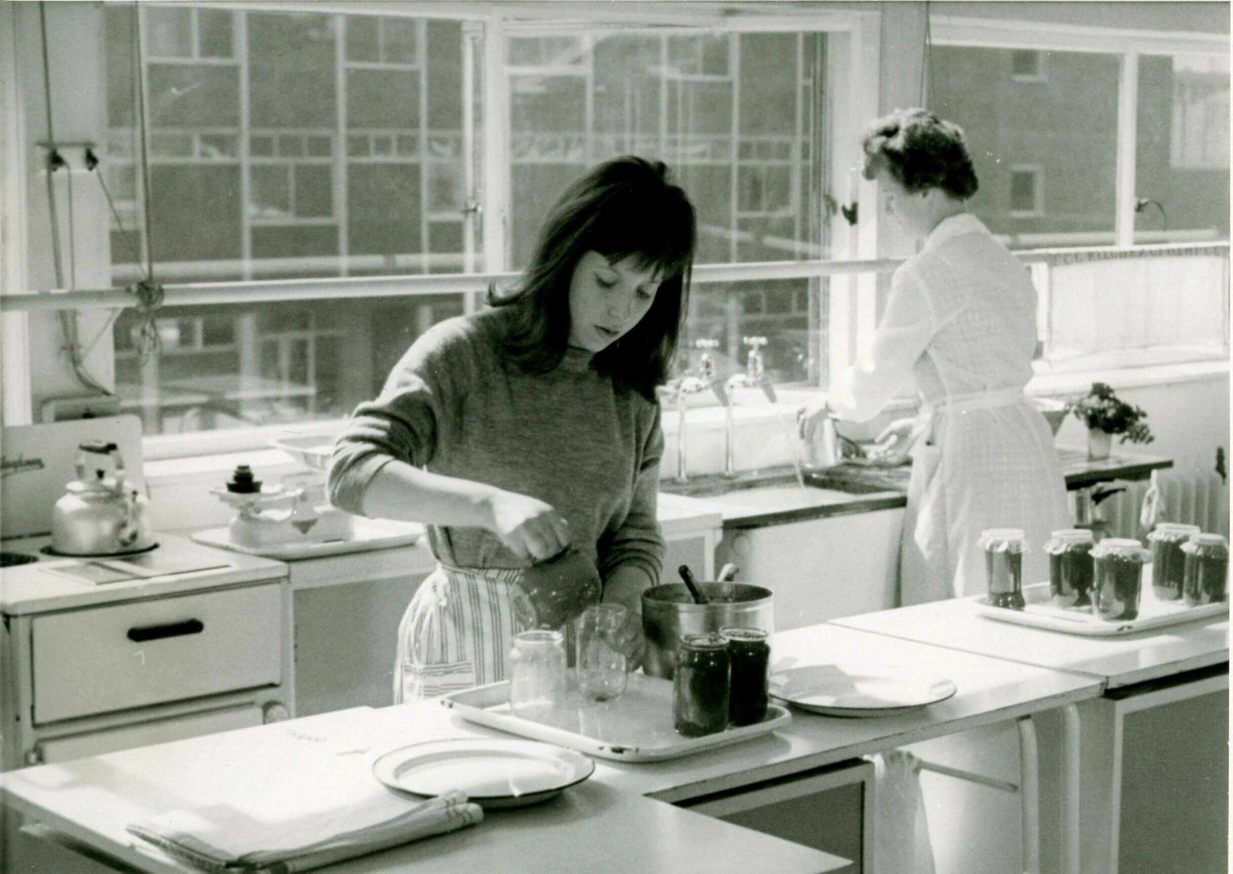 Black-and-white photograph of a kitchen with two people working at a counter. In the foreground, one person stands at a worktop stirring a mixture in a bowl, with several glass jars set out on trays nearby, some already filled. A saucepan sits on the stove to the left. In the background, another person stands at a sink beneath large windows, washing or rinsing items. The bright windows and tiled surfaces suggest a practical, well-lit workspace.