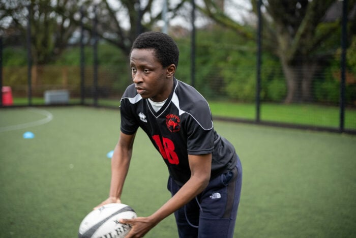 A young male athlete in a black sports jersey with the number 18 is preparing to pass a rugby ball on a grassy field. He appears focused and determined, with trees in the background and training cones scattered around the field.