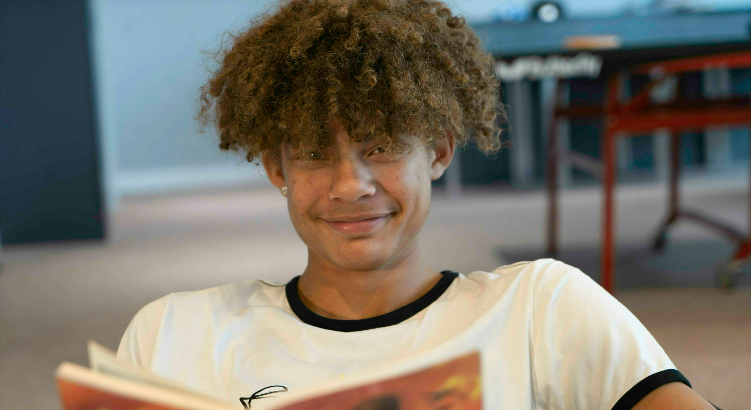 A young person sitting down holding a book and smiling, their curly hair is just covering their eyes.