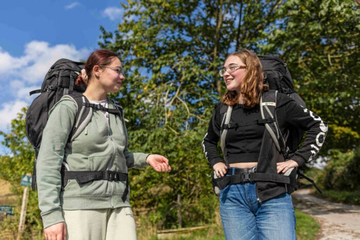 Two young women stand on a path surrounded by greenery, each wearing a large backpack. One woman, dressed in a light green zip-up hoodie and glasses, is smiling and engaging in conversation with the other, who is wearing a black long-sleeve shirt and jeans. The background features trees and a blue sky with scattered clouds.