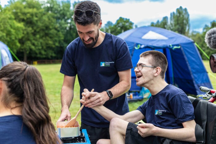 A smiling young man in a wheelchair is stirring a pot with the help of an adult man, both wearing matching navy blue t-shirts with a logo. They are outdoors in a grassy area with tents in the background. The atmosphere is cheerful and collaborative, suggesting a cooking or outdoor activity.