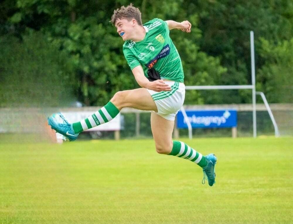 A young person in green and white football kit jumping up on the pitch after a kick.