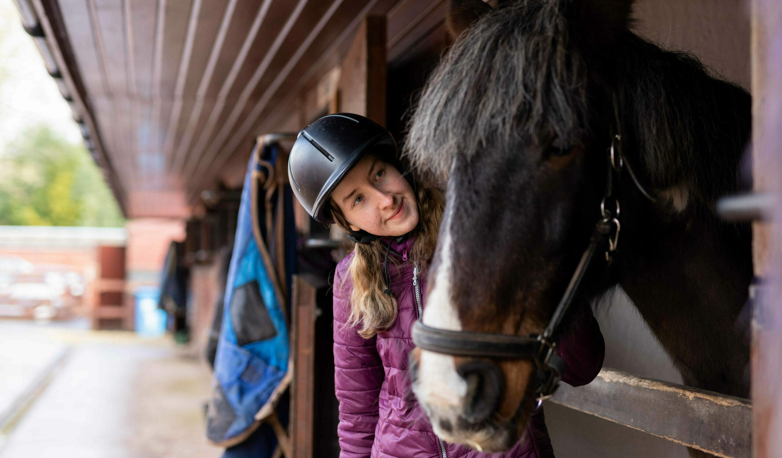 A young person wearing a riding helmet leans out from a stable and looks at a black and white horse.