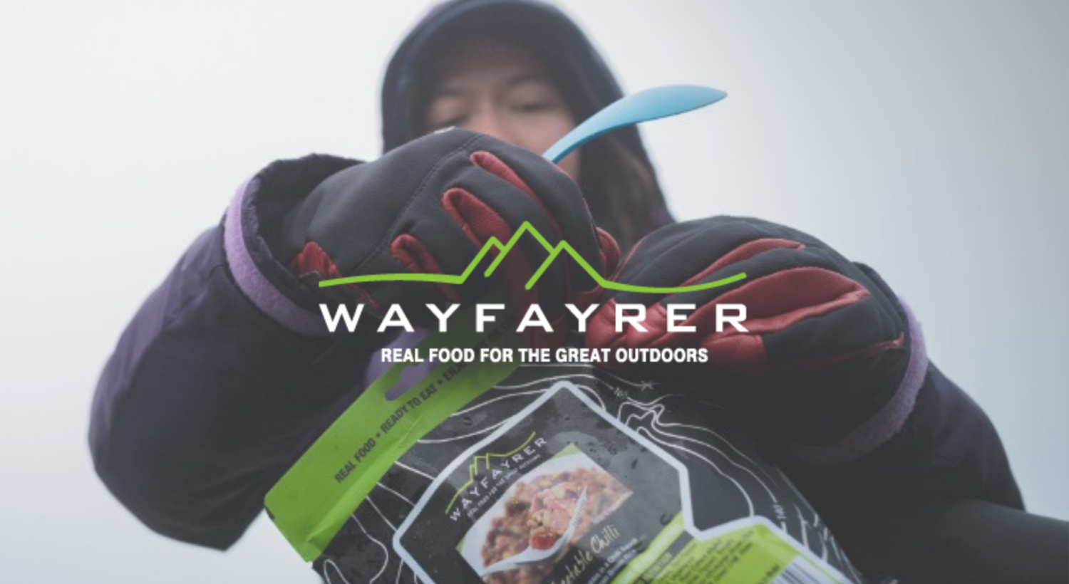 Close-up of a person outdoors wearing winter gloves and a dark jacket, holding a blue spoon and opening a Wayfayrer ready meal pouch; the Wayfayrer logo and slogan “Real Food for the Great Outdoors” are overlaid across the centre.
