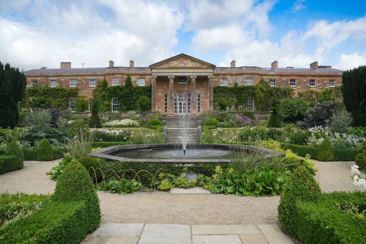 Hillsborough Castle in Northern Ireland, a large Georgian-style stone building with a central columned portico, viewed from a formal garden. A circular fountain sprays in the foreground, with steps leading up to glass doors, surrounded by neatly trimmed hedges, flower beds, and climbing plants under a partly cloudy sky.