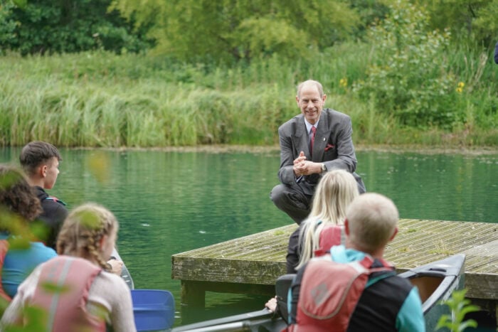 The Duke of Edinburgh crouches on the edge of a wooden jetty beside a green lake, speaking to a small group of young people. In the foreground, several young people wearing buoyancy aids sit in canoes facing the jetty. The background shows dense green trees and reeds along the water’s edge.