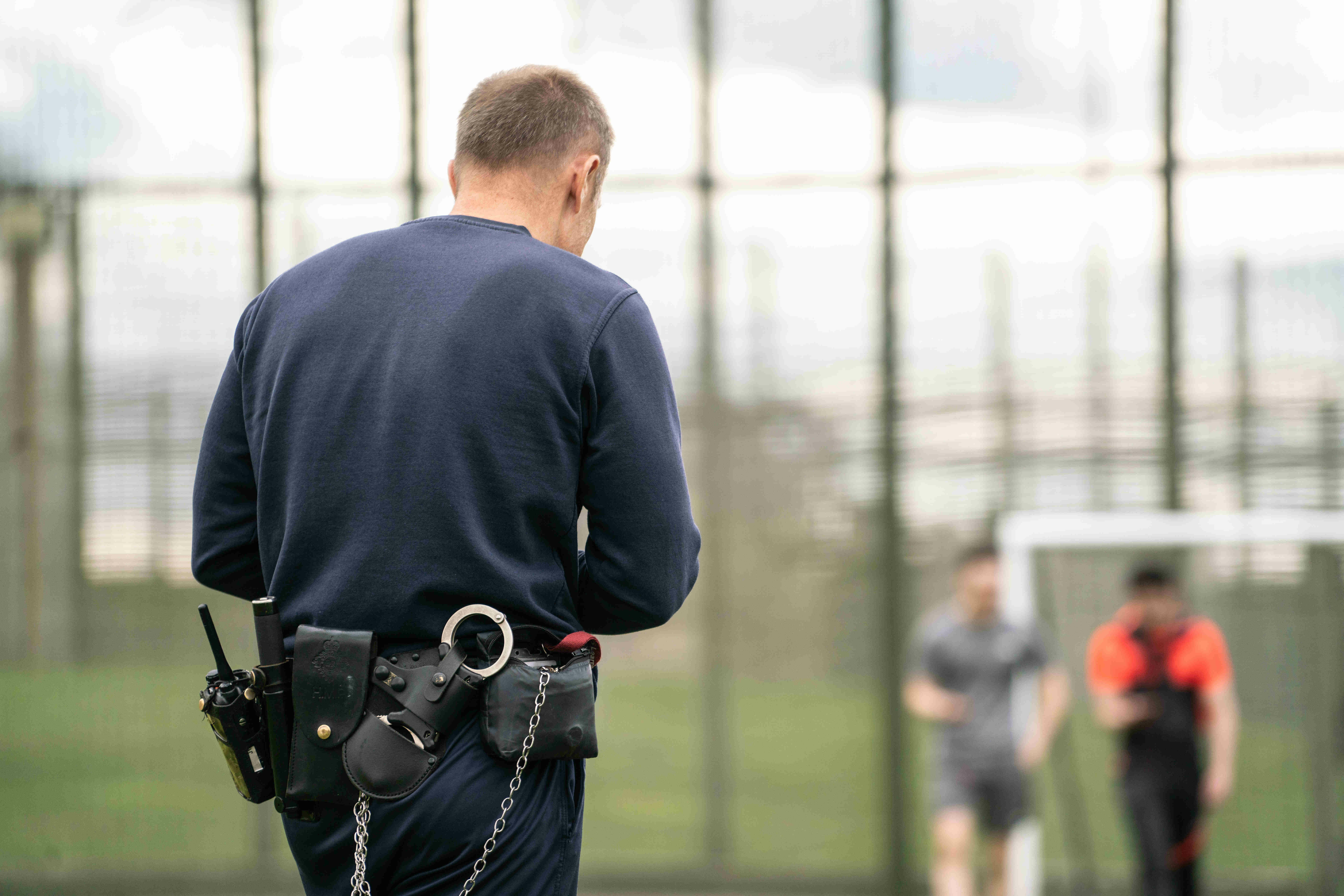 A prison officer standing on a football pitch with two young offenders in the background.