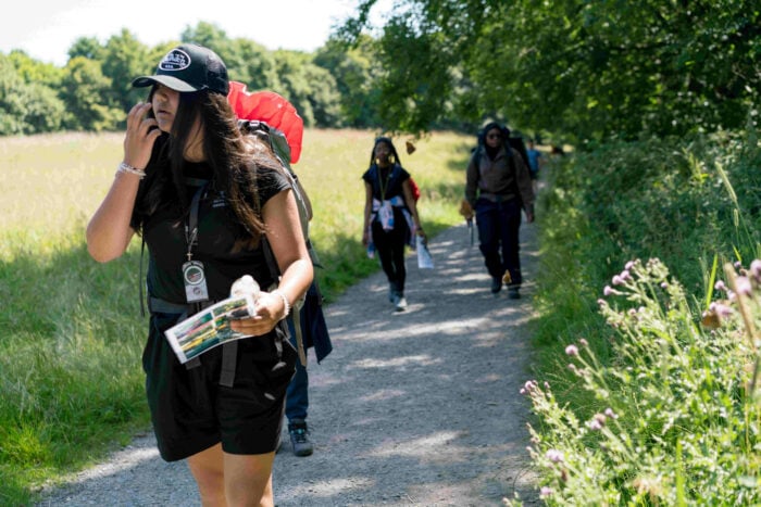 A group of people walking along a gravel path in a grassy area. The foreground features a woman wearing a black cap and a backpack, holding a map and talking on her phone. In the background, two individuals are walking, one wearing a blue jacket and the other carrying a backpack. Lush greenery surrounds the path, indicating a sunny day.