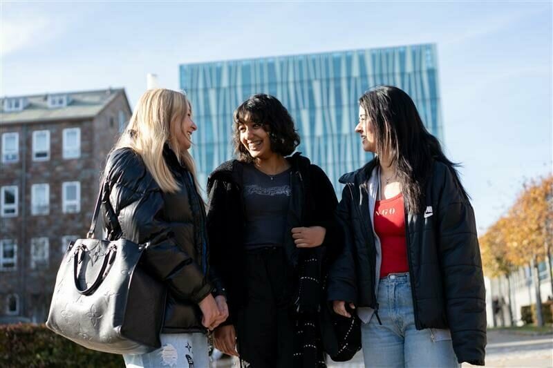 Three young people standing outdoors in an urban setting, engaged in conversation. Tall buildings are visible in the background, and the group is dressed in casual coats and jackets.