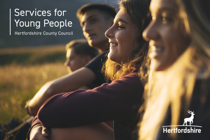 Close-up of a small group of young people outdoors in warm, golden sunlight, sitting together and looking into the distance. Overlaid text on the left reads “Services for Young People” and “Hertfordshire County Council.”