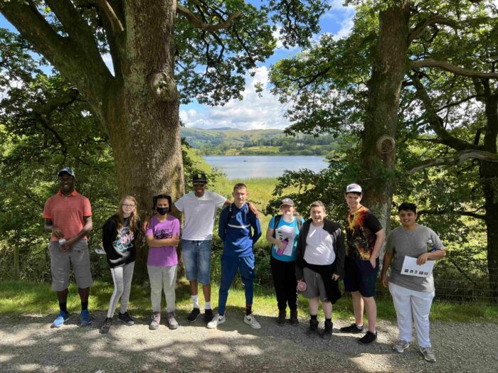 A group of ten young people stands together outdoors near a lake, surrounded by trees. The scene features a bright blue sky with fluffy clouds. The individuals are dressed in casual clothing, with some holding papers or notebooks. They appear to be enjoying a day out in nature, with a scenic view of the water and hills in the background.