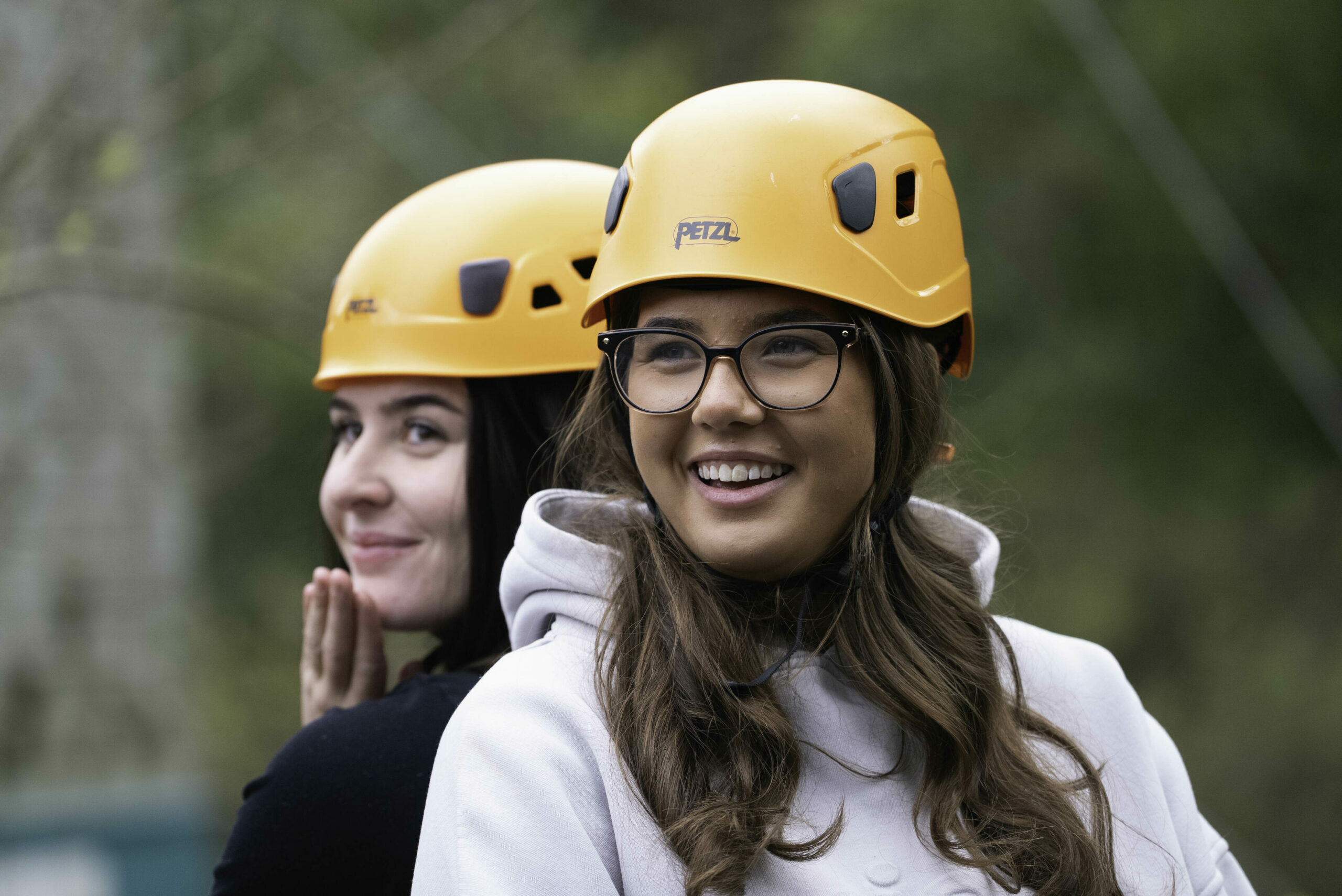 Two young women are standing back-to-back, both wearing bright yellow helmets. One woman, with long brown hair and glasses, is smiling and looking at the camera, while the other woman, with dark hair, is playfully posing with her hand on her cheek. They are outdoors, surrounded by greenery.
