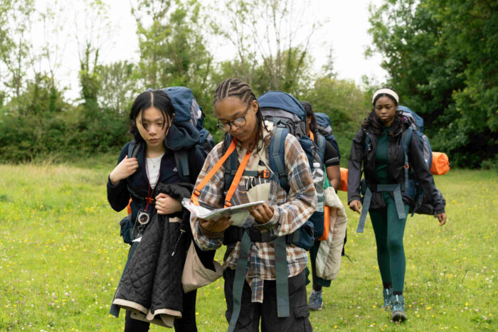 Four young people walking through a grassy field carrying rucksacks and reading a map.