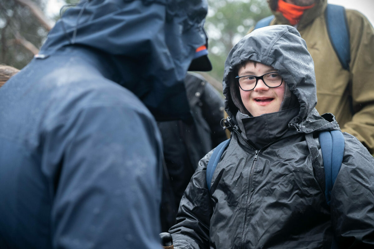A young person with glasses and a joyful expression is wearing a black rain jacket and hood, standing outdoors in rainy weather.