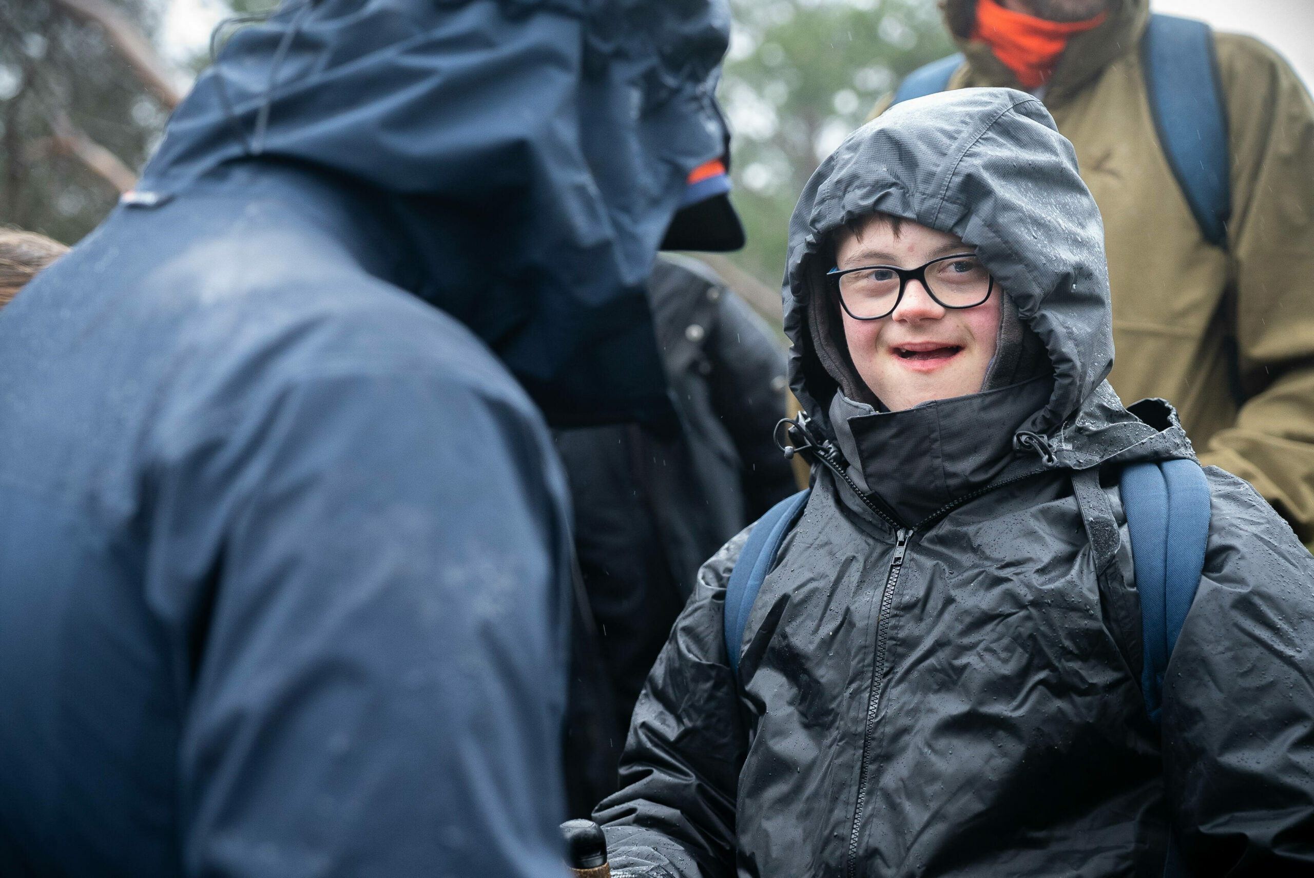 A young person with glasses and a joyful expression is wearing a black rain jacket and hood, standing outdoors in rainy weather.