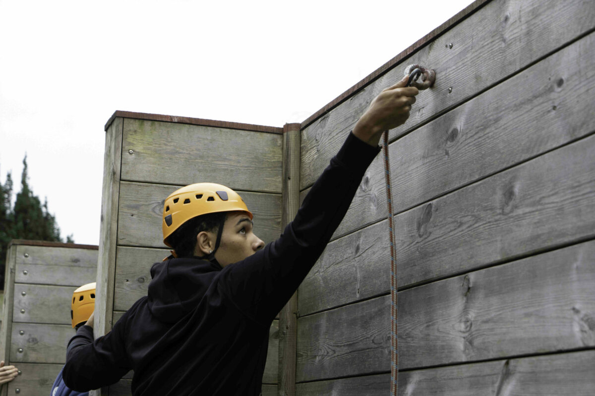 A person wearing a yellow helmet reaches up to secure a climbing rope to a wooden wall. The individual is focused and appears to be participating in an outdoor climbing or obstacle course activity. Another person is partially visible in the background, also engaged in the activity. The setting is outdoors, with trees visible in the distance.