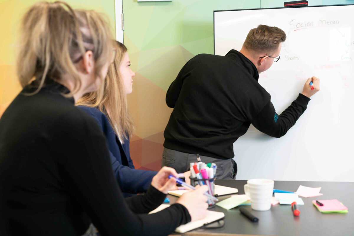 A group of three people in a meeting room. Two individuals are seated at a table, one with long hair and the other with medium-length hair, both looking towards a person standing and writing on a whiteboard. The standing person is wearing a black sweater and is focused on writing notes related to 