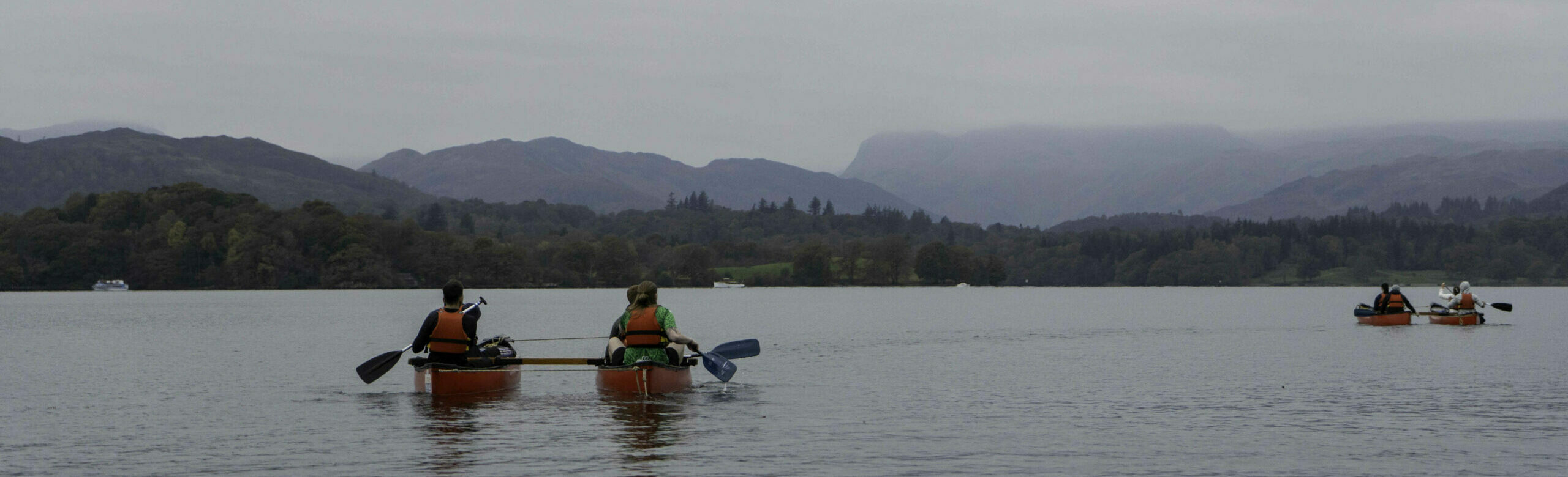 Two kayakers paddle on a calm lake surrounded by misty mountains and overcast skies. The landscape features lush green hills in the background, creating a serene outdoor scene.