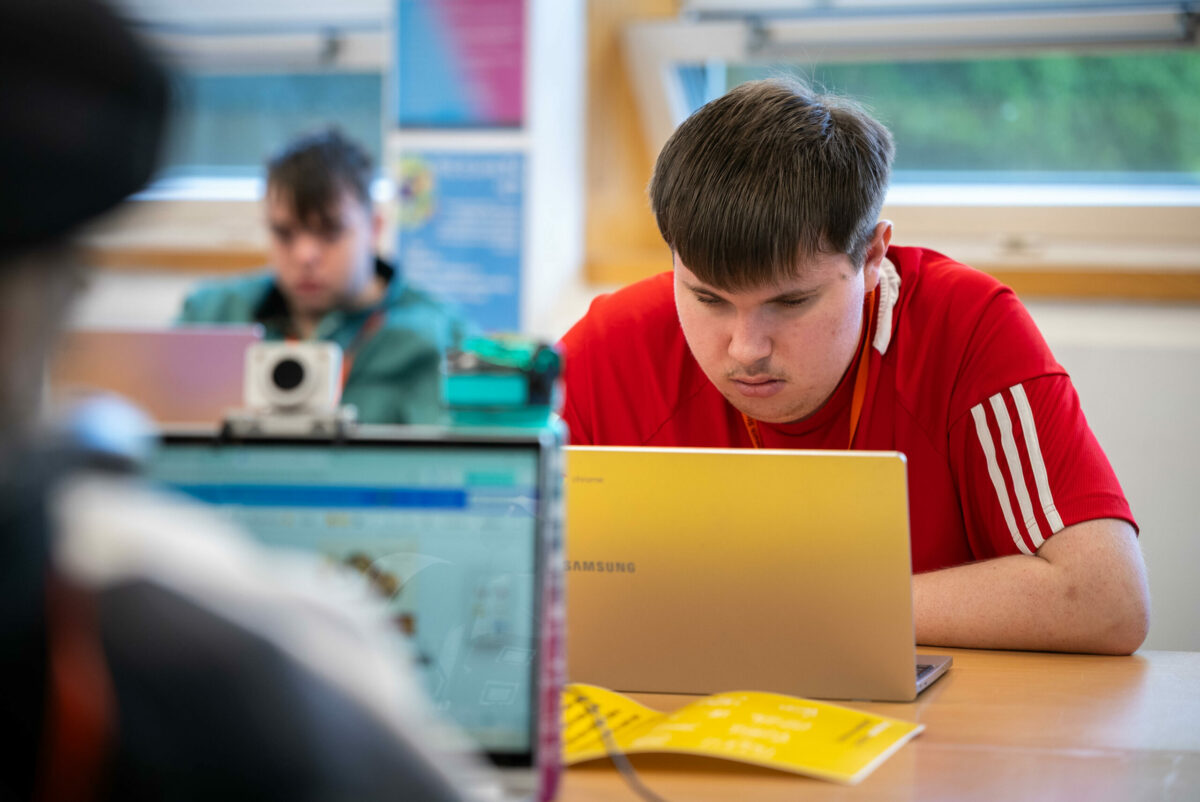 A young person in a red tshirt leaning over their laptop in a classroom.