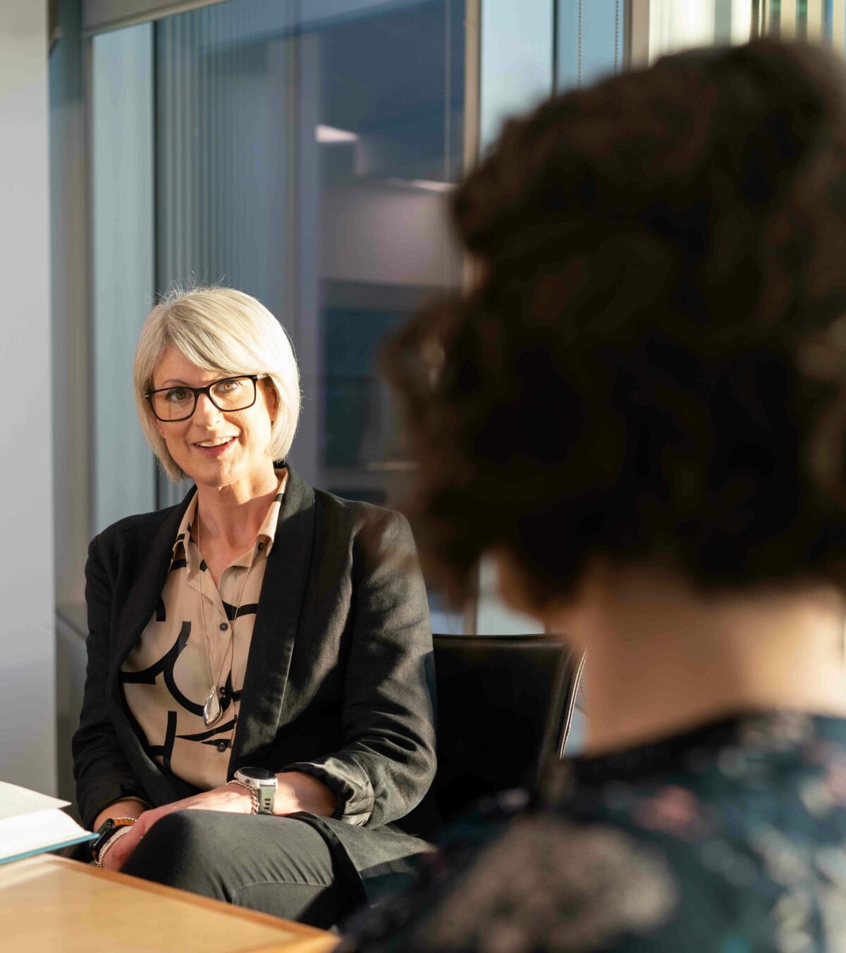 A woman with short, light-colored hair and glasses sitting at a table, smiling and engaging in conversation.
