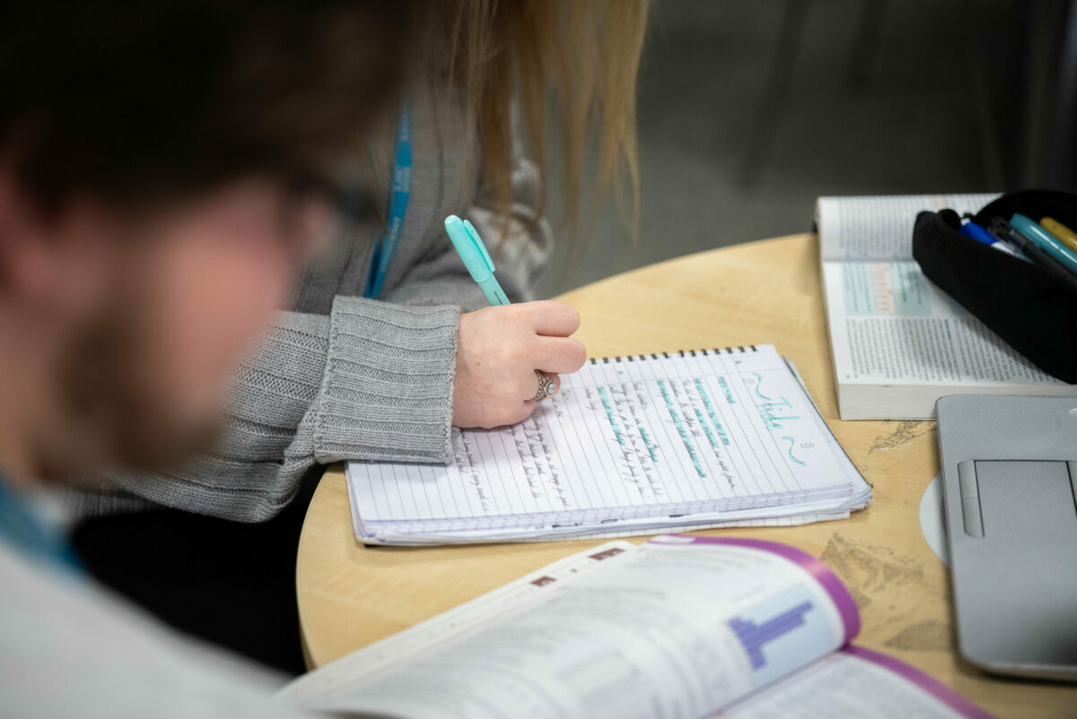 A person's hand writing in a notebook with a turquoise pen.