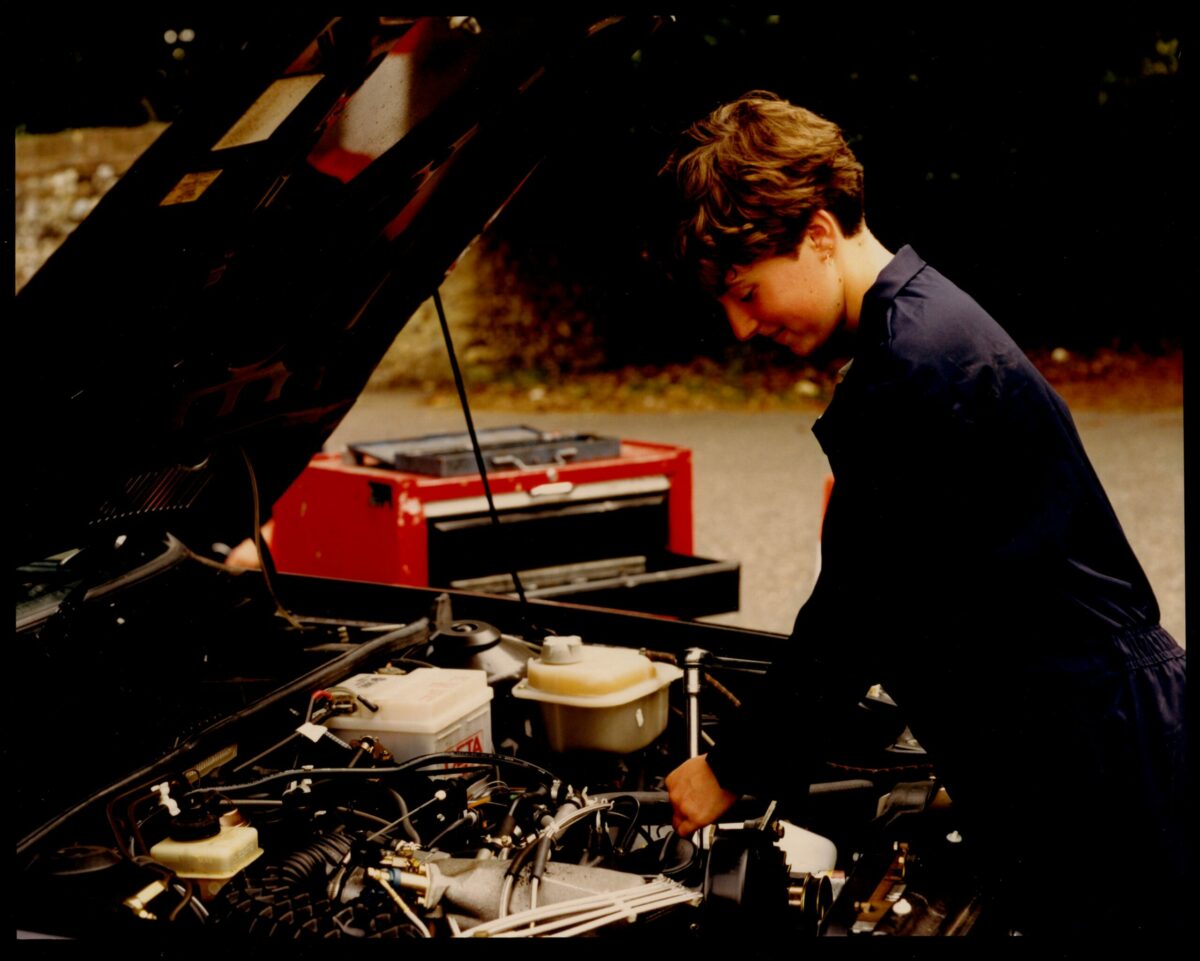A young person in a dark blue jumpsuit is working on the engine of a car with the hood raised.