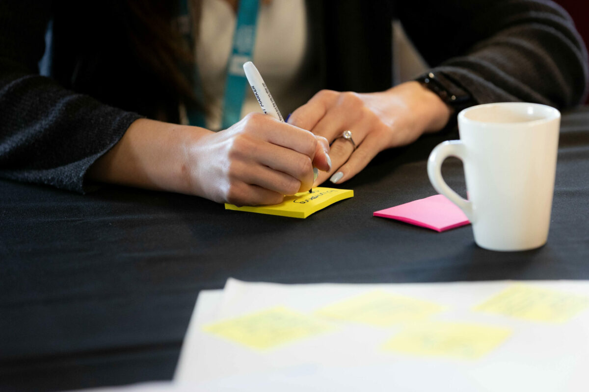A close-up of a person's hand writing on a yellow sticky note with a black marker.