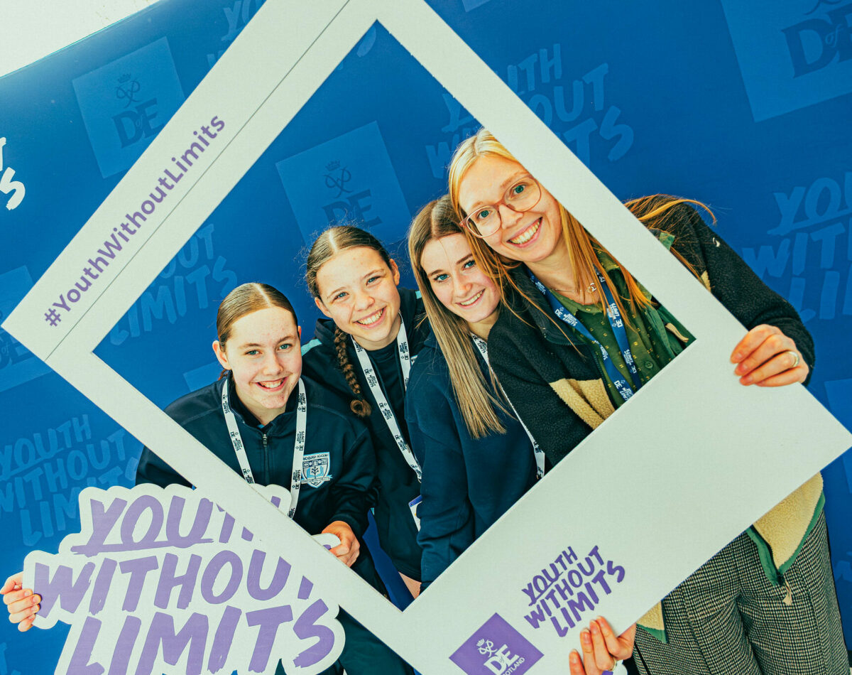 Four attendees pose in front of a blue Youth Without Limits and DofE Scotland backdrop, holding a large photo-frame prop branded with “#YouthWithoutLimits