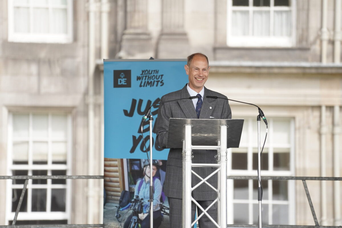 HRH The Duke of Edinburgh stands at a lectern on an outdoor stage in the Gardens of the Palace of Holyroodhouse delivering a speech.