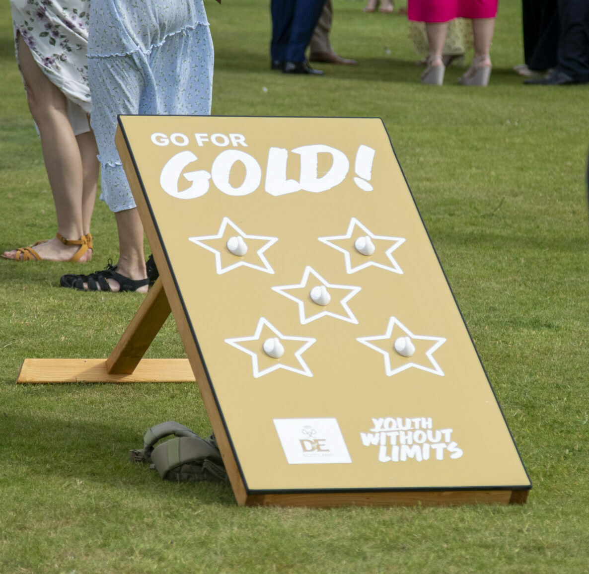 A gold-coloured DofE-branded lawn game board stands on a grassy outdoor event space. The board displays the text “GO FOR GOLD!” above five star-shaped targets and includes DofE and Youth Without Limits logos. Groups of attendees are gathered and talking in the background, while a person walks past in the foreground carrying a tambourine. The image captures a social outdoor celebration with activities taking place on a lawn.