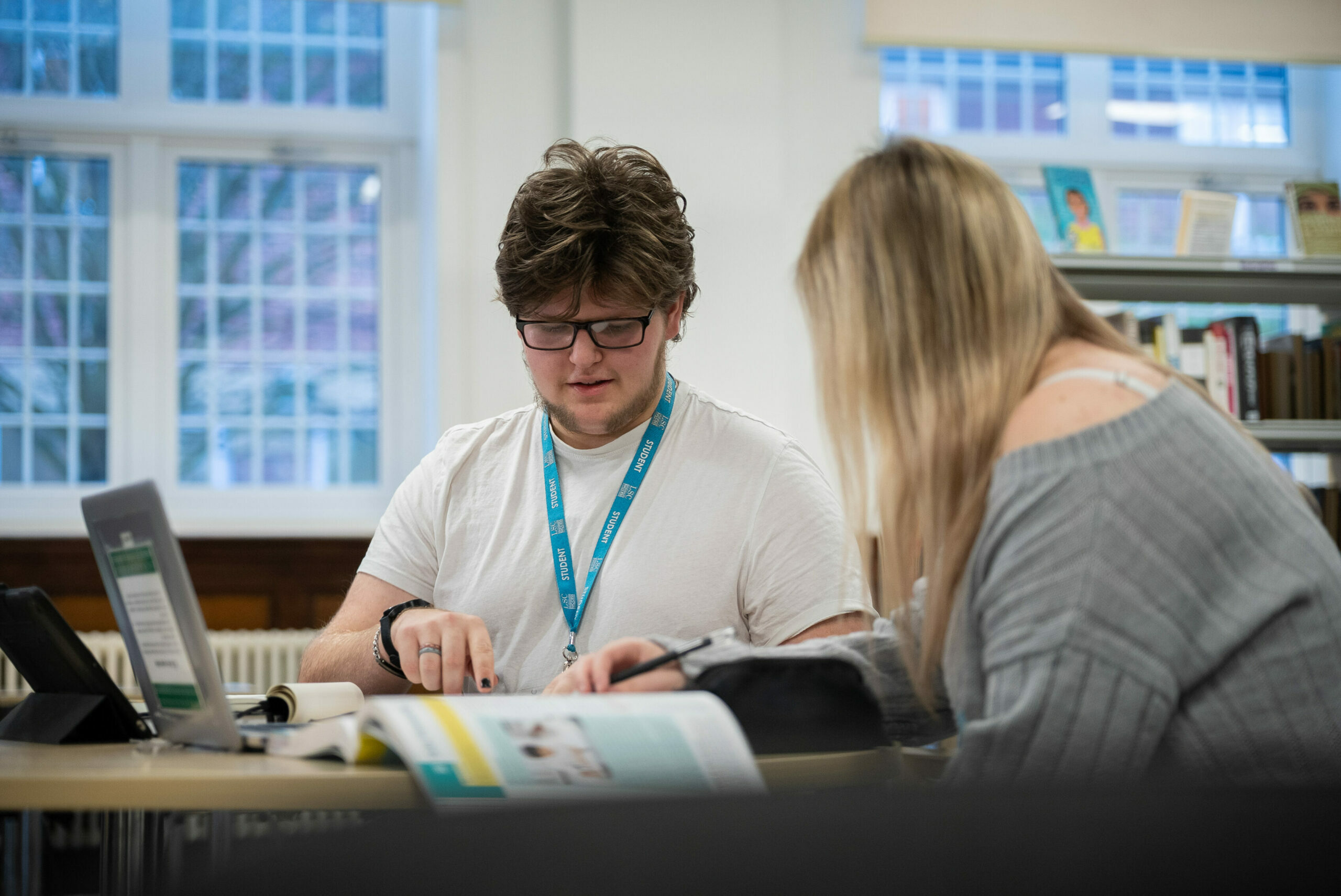 A young man with glasses and a white t-shirt is sitting at a table in a library, focused on a book. He is pointing at something in the book while discussing it with a young woman beside him, who has long blonde hair and is wearing a gray sweater. A laptop and other materials are on the table, and large windows in the background let in natural light.