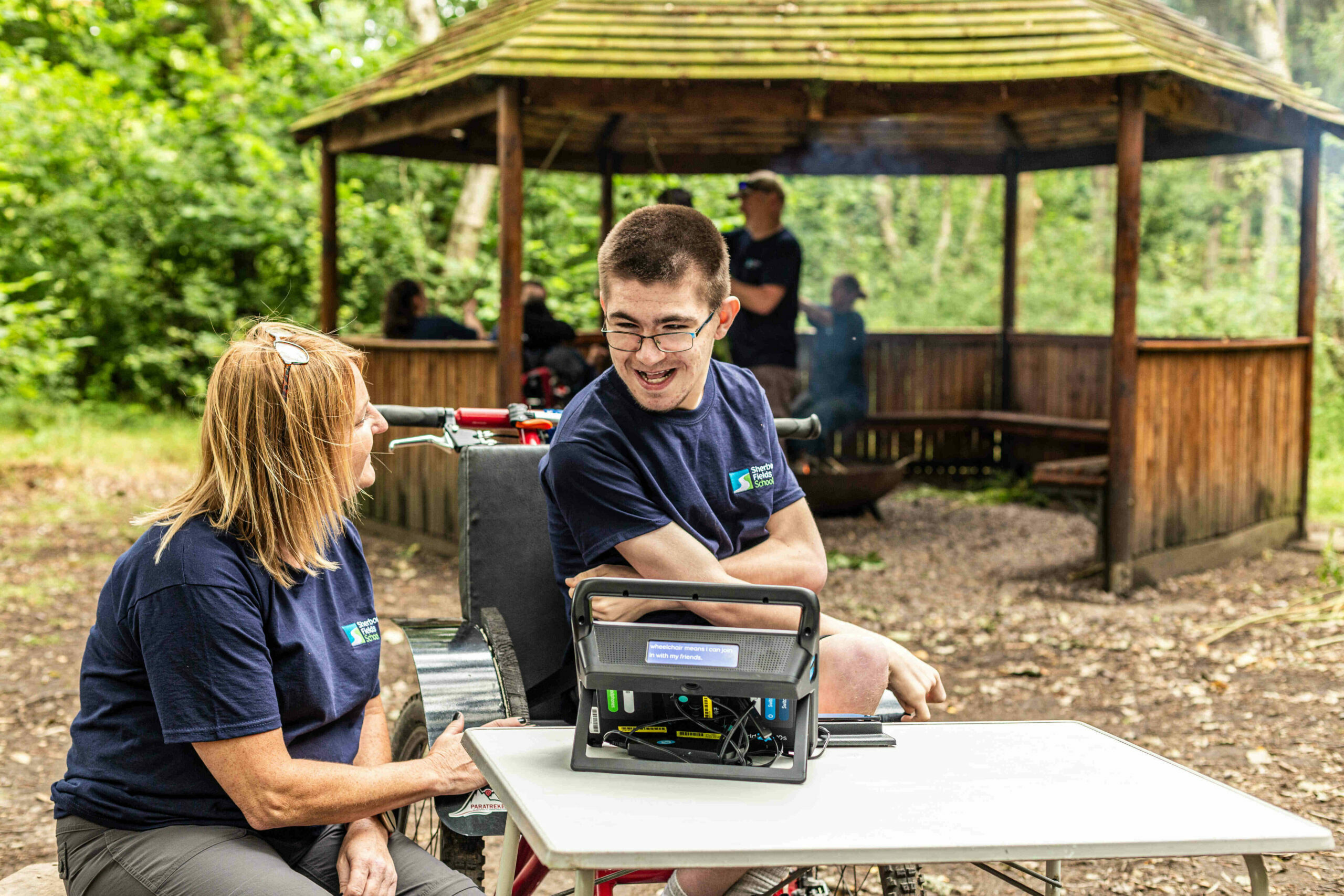 A young person in a wheelchair smiling at an adult sitting on a bench in a forest.